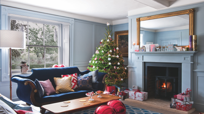 Pastel blue living room, moulded wood wall panelling, fireplace lit wood burning stove, Christmas tree, presents, blue vintage antique sofa, assorted cushions, giant red baubles, gilded framed mirror