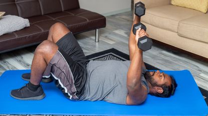 A man performs a chest press at home with dumbbells. He is lying on an exercise mat, with his knees bent and feet flat on the floor. He holds a pair of dumbbells above his head, with his arms straight. Behind him we see two couches.