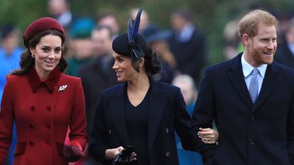 Kate Middleton wears a red coat with a fur collar and a matching hat, Meghan Markle wears a navy coat and matching dress, Prince Harry wears a white shirt, blue tie, and large overcoat