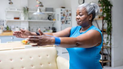 A woman exercises with a resistance band inside a house. She is standing with her arms out in front of her and a short looped resistance band around her wrists. Her hair is tied back and she is wearing a t-shirt. Behind her we see a couch, shelving and a decorated fridge. 