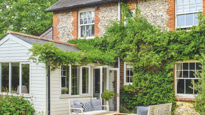 A brick and flint house with a large wisteria, patio with grey wooden benches and a fire pit beside the lawn.