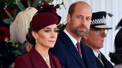 Kate Middleton wearing a burgundy coat and hat standing next to Prince William wearing a blue coat and red tie, both with thoughtful expressions