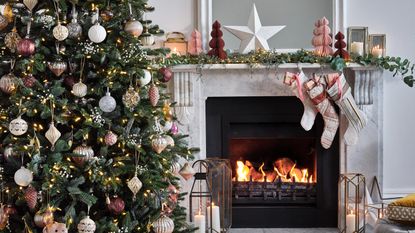 A decorated Christmas tree with pink, gold and burgundy baubles set next to a marble fireplace decorated with an eucalyptus garland and Christmas stockings