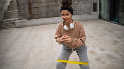 Woman outside with yellow resistance band wrapped above her knees. She wears a sweatshirt and sweatpants. 