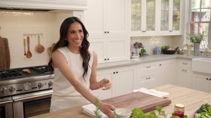 Meghan Markle in the kitchen smiling and wearing a beige outfit