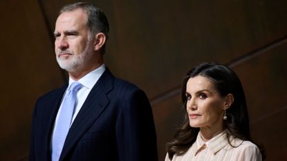 King Felipe of Spain wearing a blue suit and tie standing next to Queen Letizia wearing a peach dress with a collar in front of a brown wall