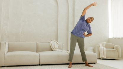 Senior woman reaches her right arm above her and to the left. She stands in front of a white couch 