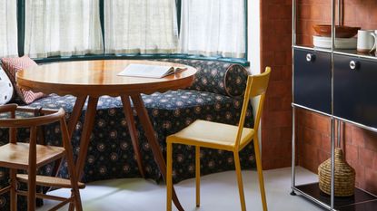 upholstered banquette seat in dining room with terracotta tiles, yellow chair, and timber table