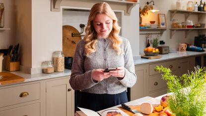 Blonde woman wearing grey wool cardigan looking at phone in her hands in a beige kitchen with white worktops and interior wall lights