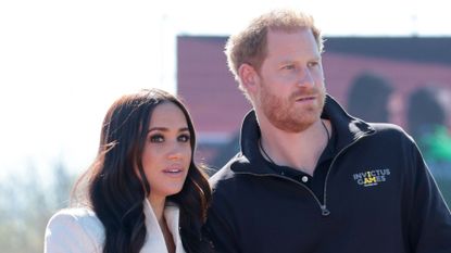 Meghan Markle has long brown hair and is wearing a white jacket while she stands with husband Prince Harry, who wears a black polo shirt with the Invictus Games logo