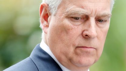 A closeup picture of Prince Andrew looking upset and wearing a blue suit with a white dress shirt and red tie