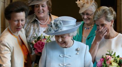 Queen Elizabeth wears a light blue suit and matching hat and everyone laughs as she cuts a cake