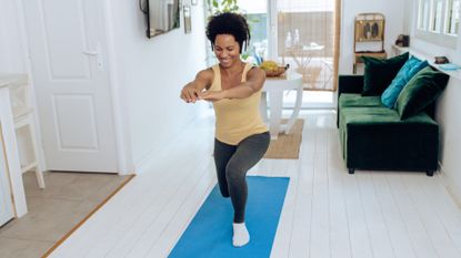 woman wearing yellow vest, grey leggings and headphones doing a forward lunge on a blue mat. she&#039;s in a white living-home setting with a dark green sofa behind her 