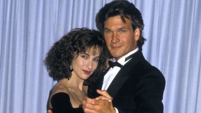 Jennifer Grey wears a black dress while holding a dance pose with Patrick Swayze, her Dirty Dancing co-star who is wearing a tuxedo, as they stand in front of an Oscar statue