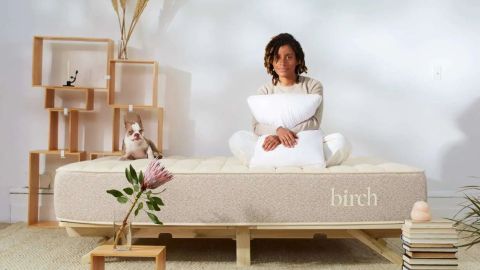 The image shows a woman sitting on the Birch Natural mattress on a wooden bed frame in a neutrally decorated bedroom