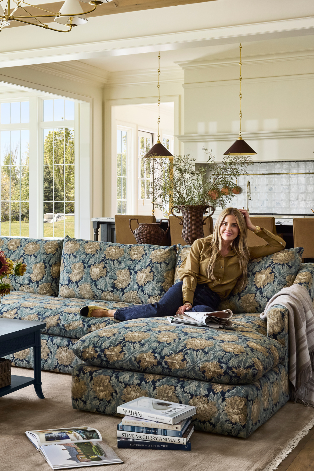 Shea McGee smiles on a floral jacquard sectional from her new McGee &amp;amp; Co. fall 2026 collection, which is styled with a navy storage coffee table, taupe rug, woven table lamp, and collected objects
