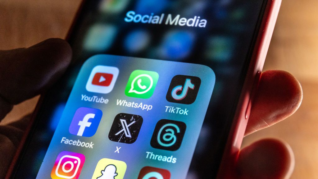 A man holds a smartphone iPhone screen showing various social media apps including YouTube, TikTok, Facebook, Threads, Instagram and X