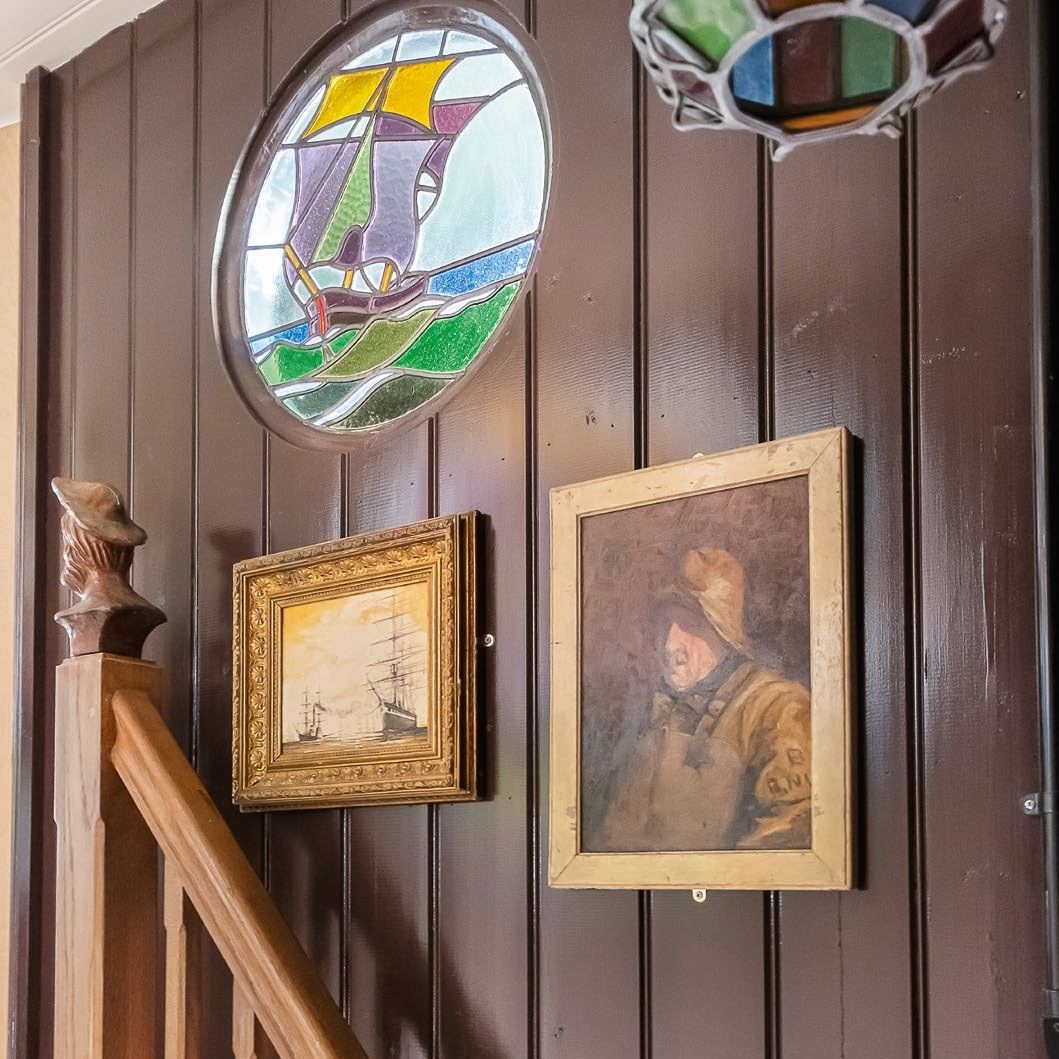 View of a dark brown wood panelled wall beside a staircase with small round stained glass window and antique nautical artwork