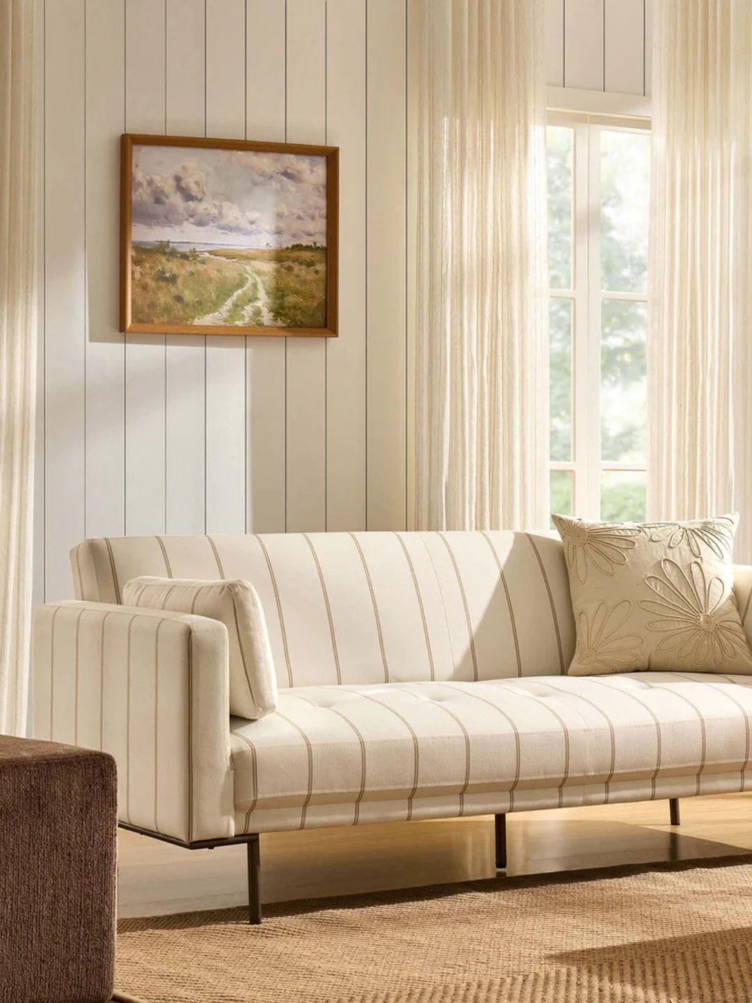 A cream striped sofa bed in a living room with cream striped walls and cream curtains with a wood-framed picture of the countryside in the middle of the wall behind