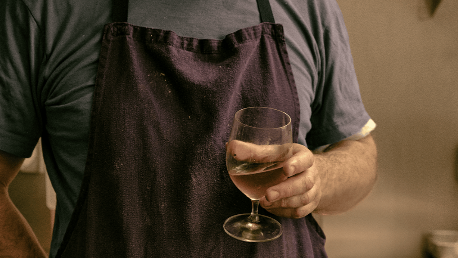 a film camera image of a man holding a glass of ros&amp;eacute; whilst wearing a navy blue apron over a blue teeshirt