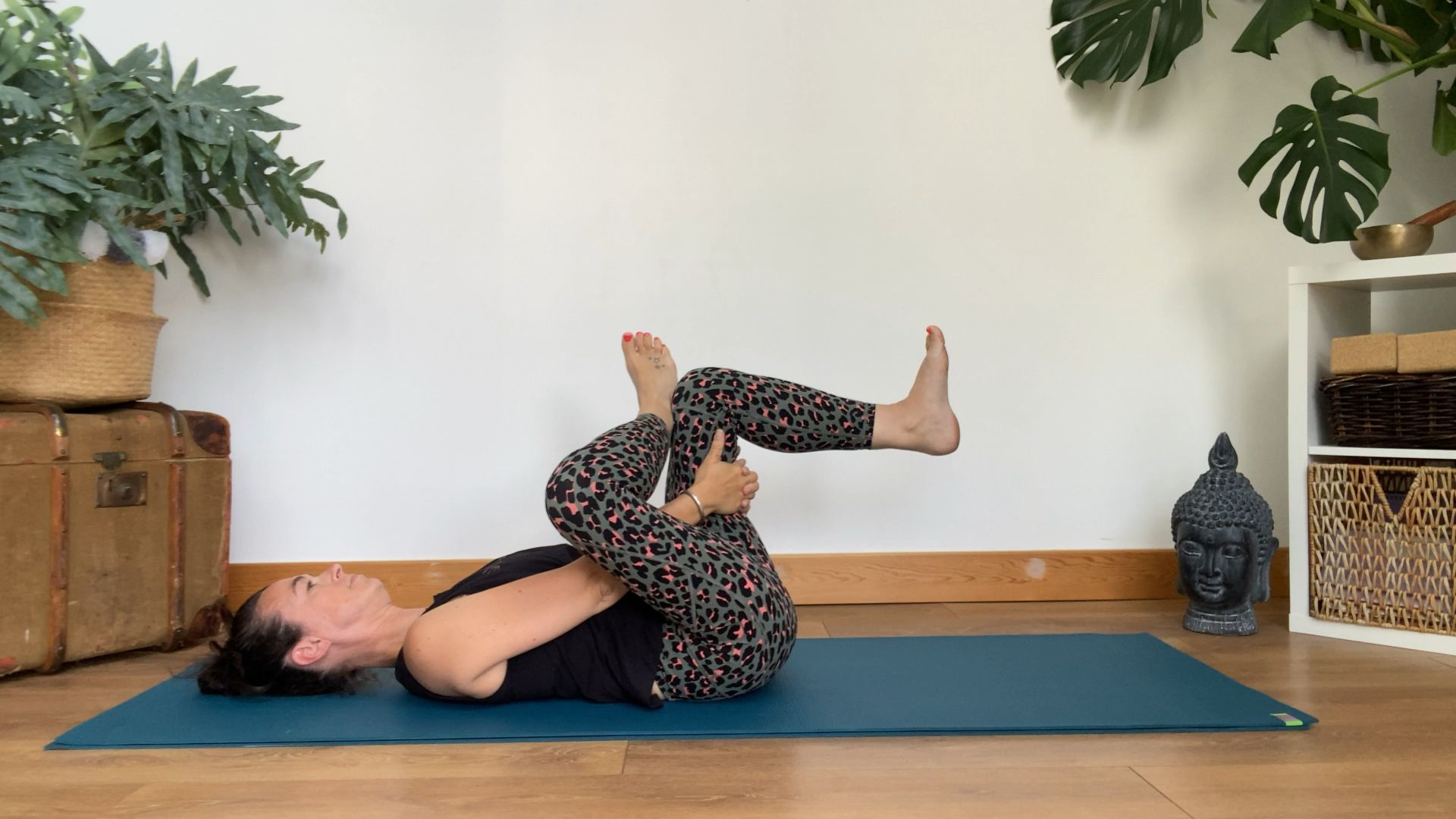 Louise Bartlett lies on her back in her living room performing a figure four stretch on a yoga mat. Her left knee is bent, her thigh pointing upwards, shin parallel to the floor. Her right knee is also bent, with the ankle crossed over the left thigh. Her hands are looped around her left thigh.