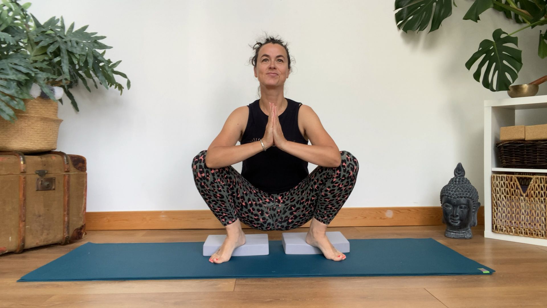 Louise Bartlett perform a deep squat at home, with her heels elevated on yoga blocks and her hands together at her chest.