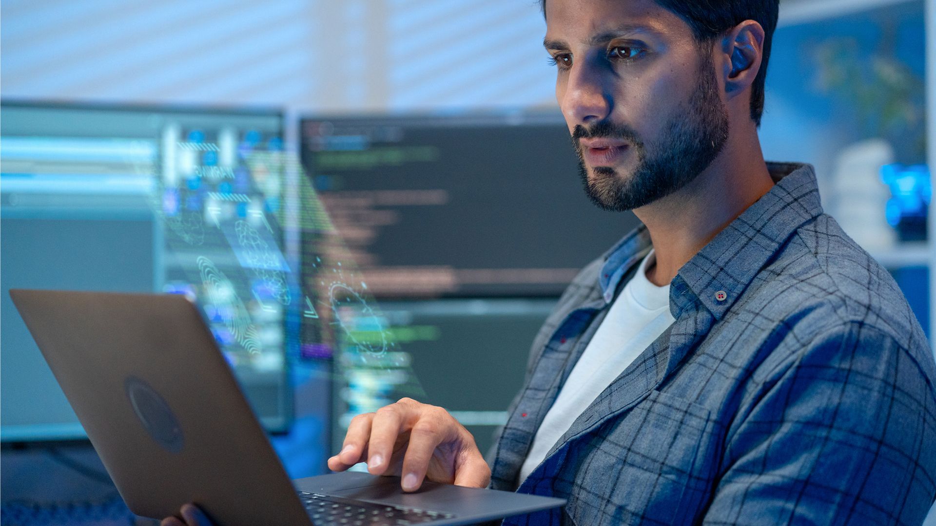 A man stands in front of several monitors, holding up a laptop