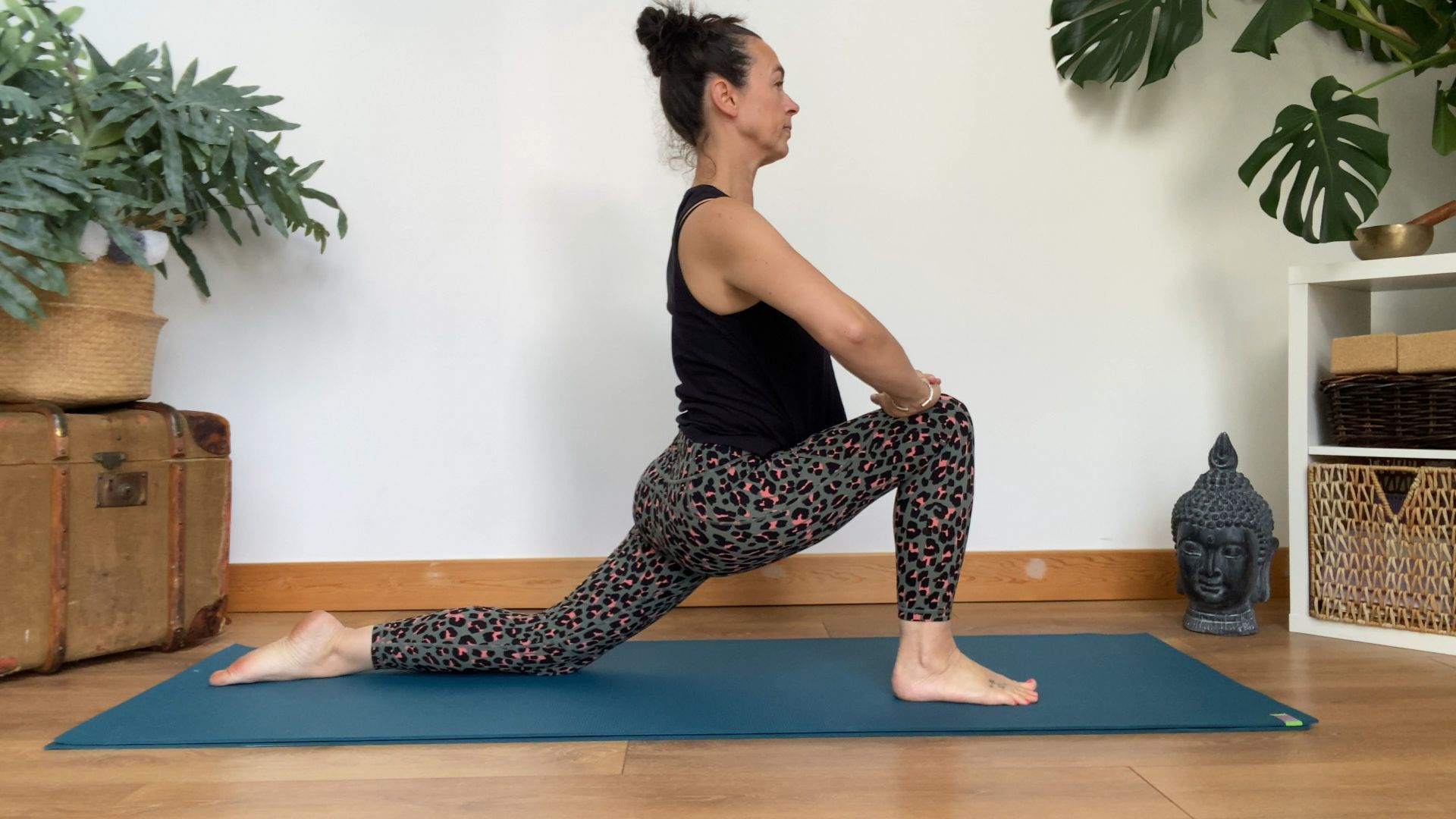 Louise Bartlett performs a low lunge at home on a yoga mat. We see a trunk, leafy plants, bookshelves and a buddha hear in the background.