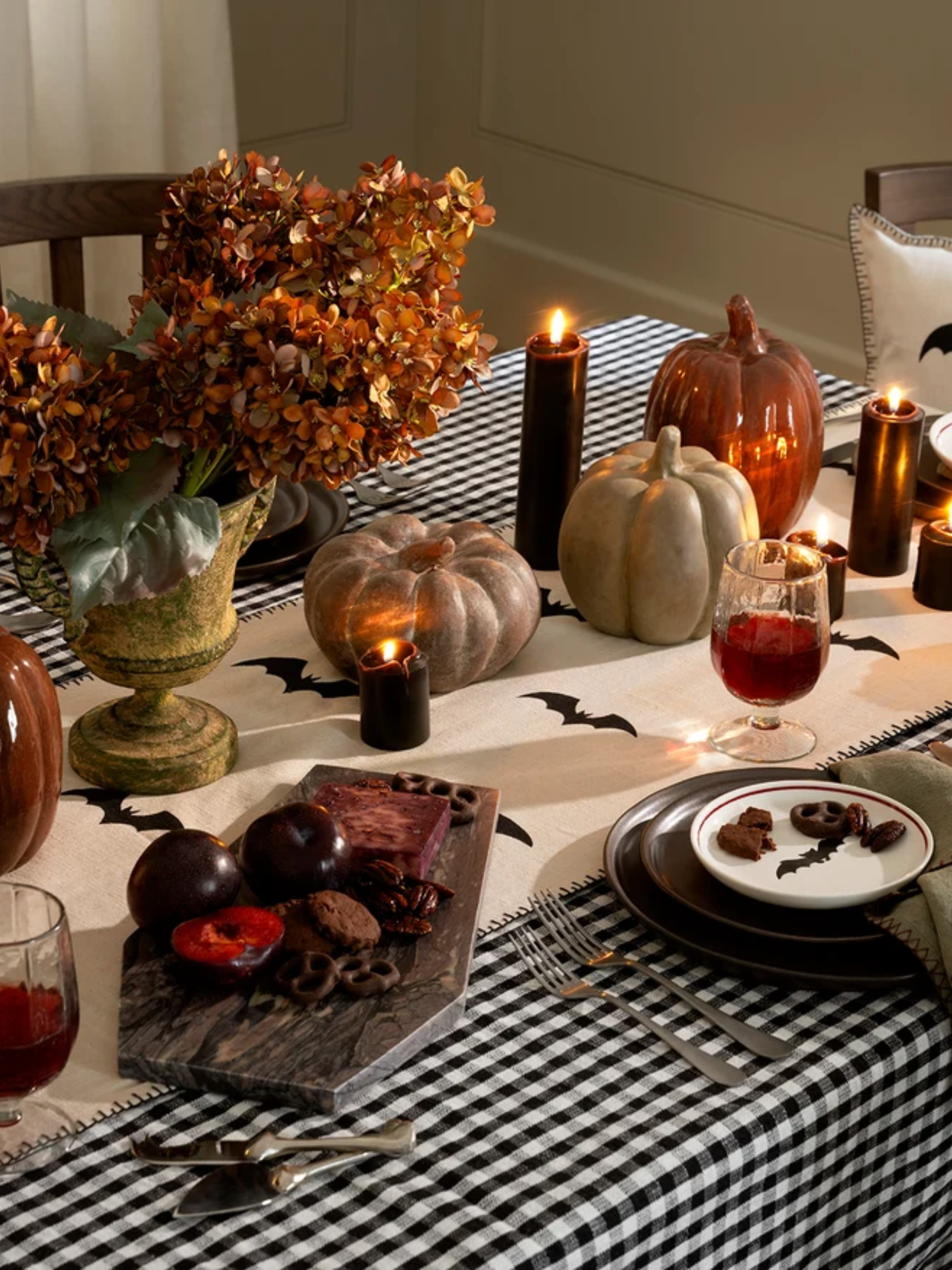 Timeless, richly layered Halloween tablespace featuring a black-and-white gingham tablecloth, marble coffin-shaped cheese board, stacks of chocolate brown plates. and black taper candles aglow.