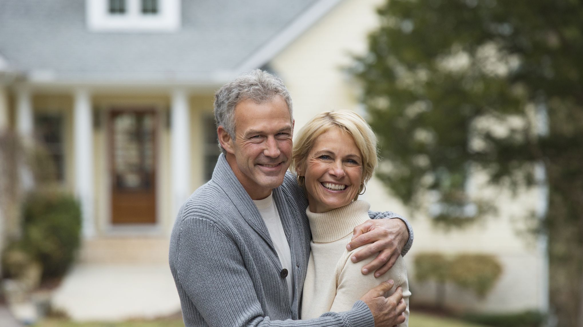A wealthy couple hugs in front of their house.