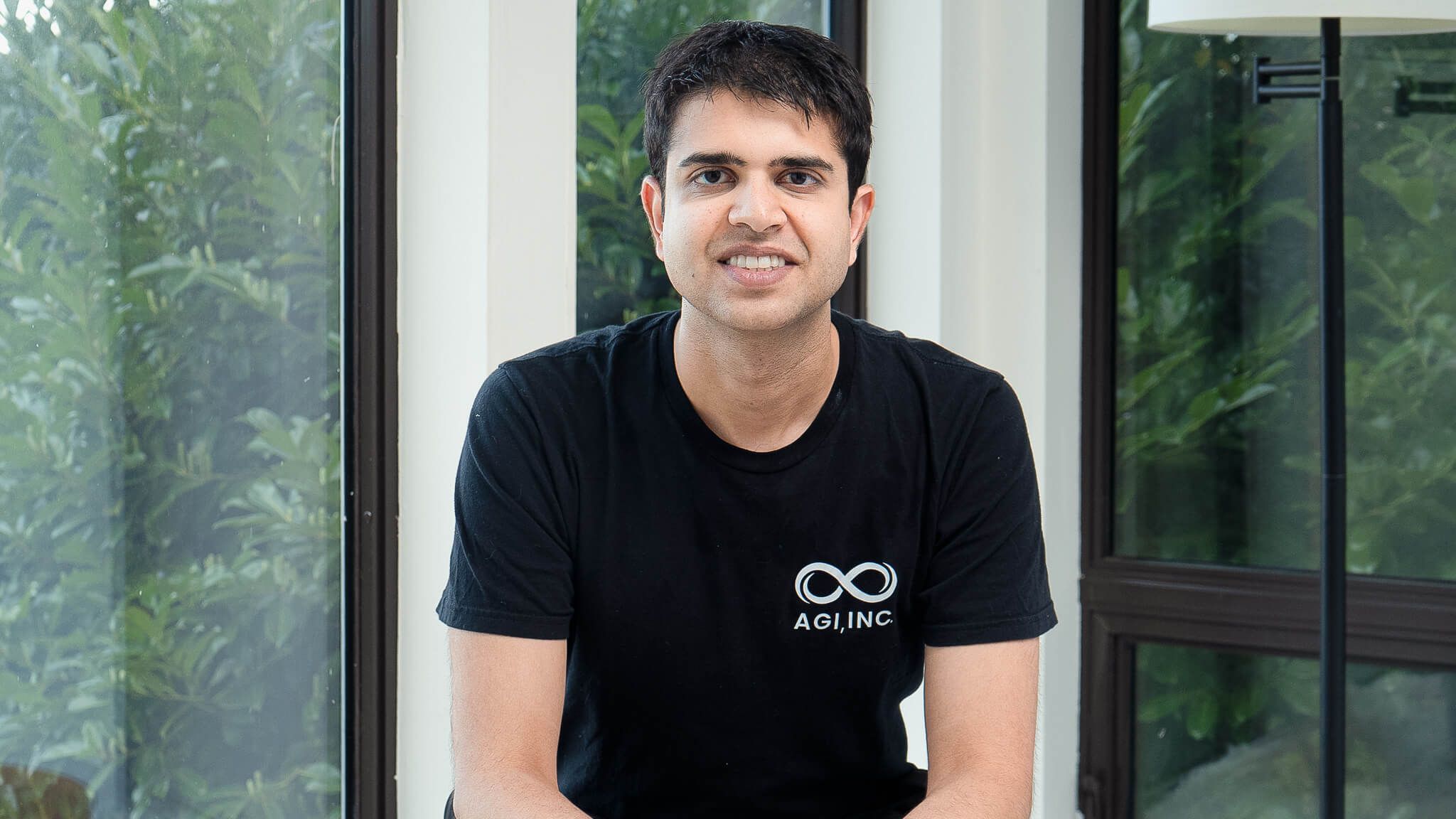 A photograph of Div Garg, smiling, wearing a black AGI T-shirt, sitting in front of a window with green trees outside
