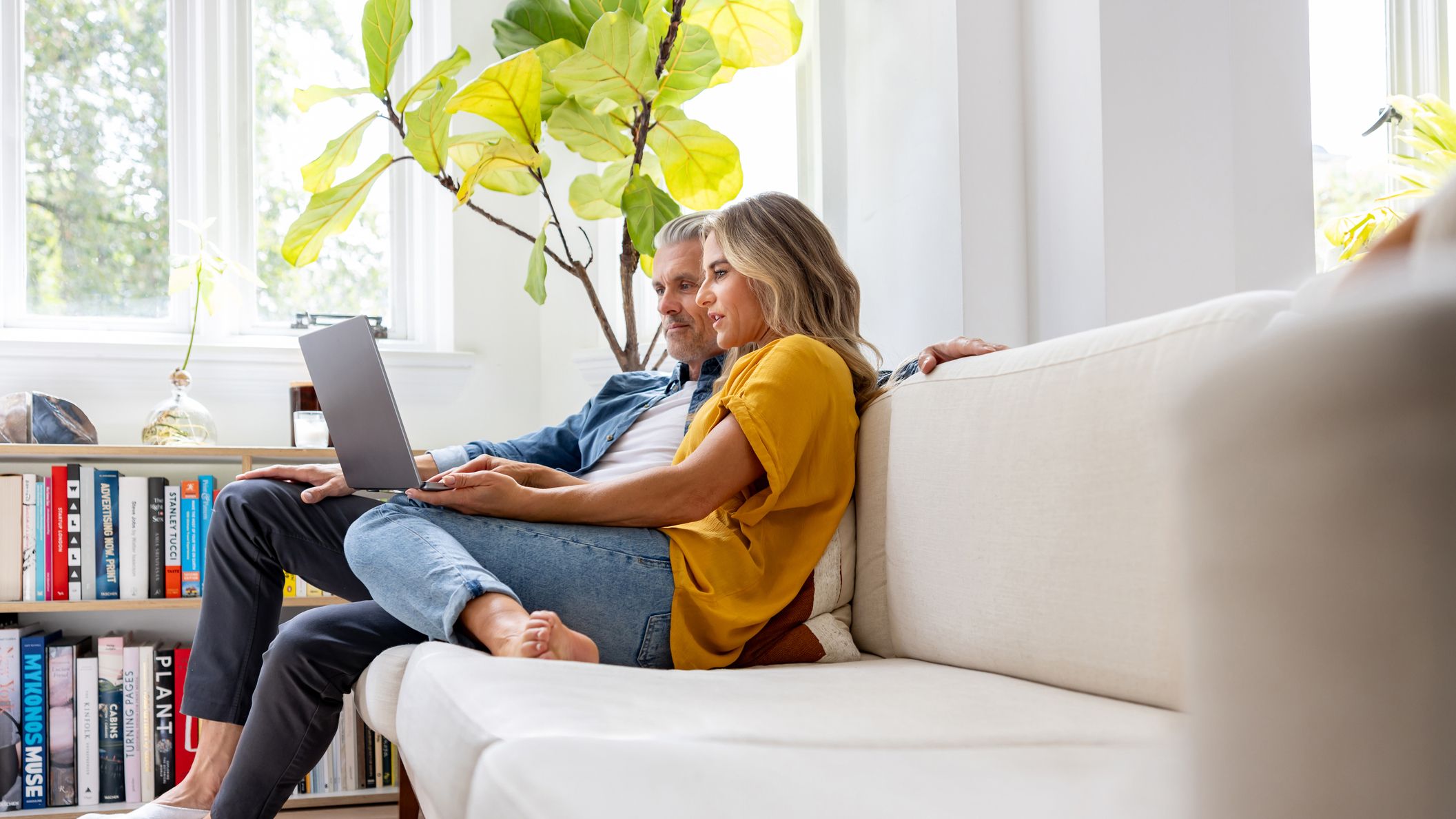 A couple in their late 50s or early 60s relaxes on their couch at home. They are looking at a laptop together and seem to be concentrating.