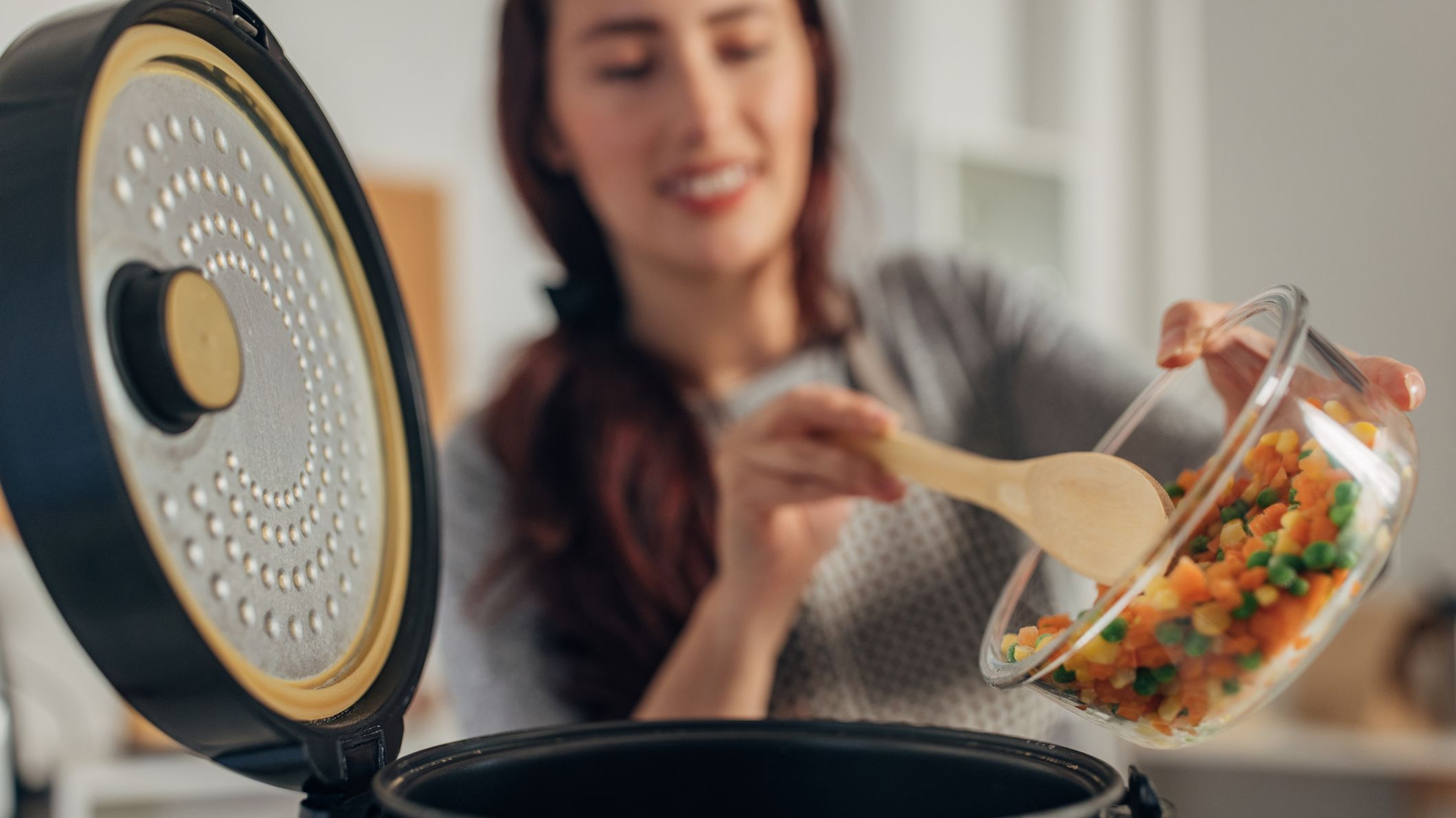 Woman using a wooden spoon to empty a glass bowl of chopped veg into slow cooker