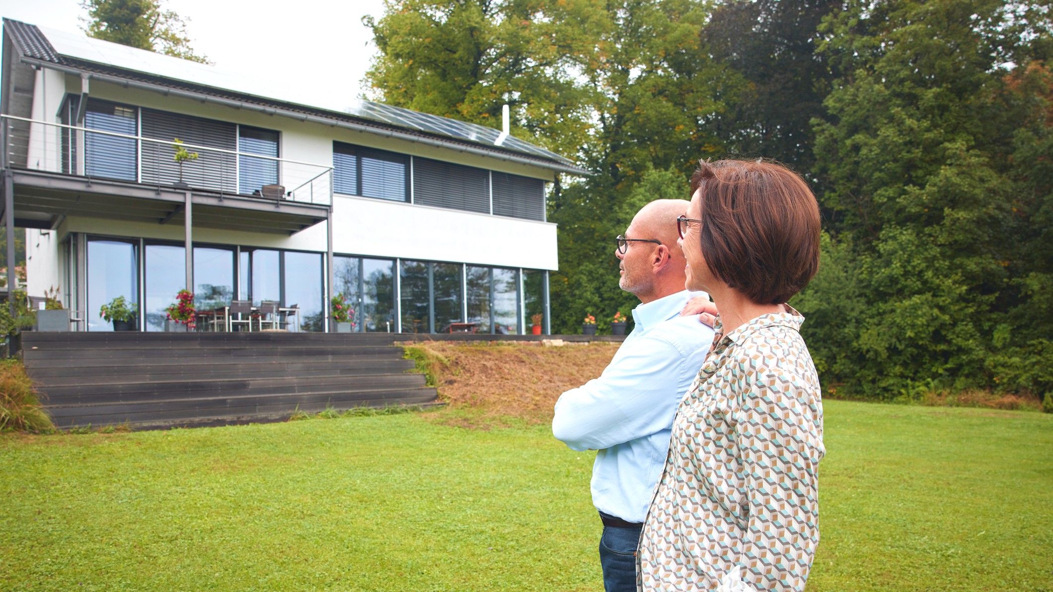 An older couple stands in their backyard, looking at their modern home. There are steps leading up to the porch with no bannister, indicating the home may not be ideal for aging in place.