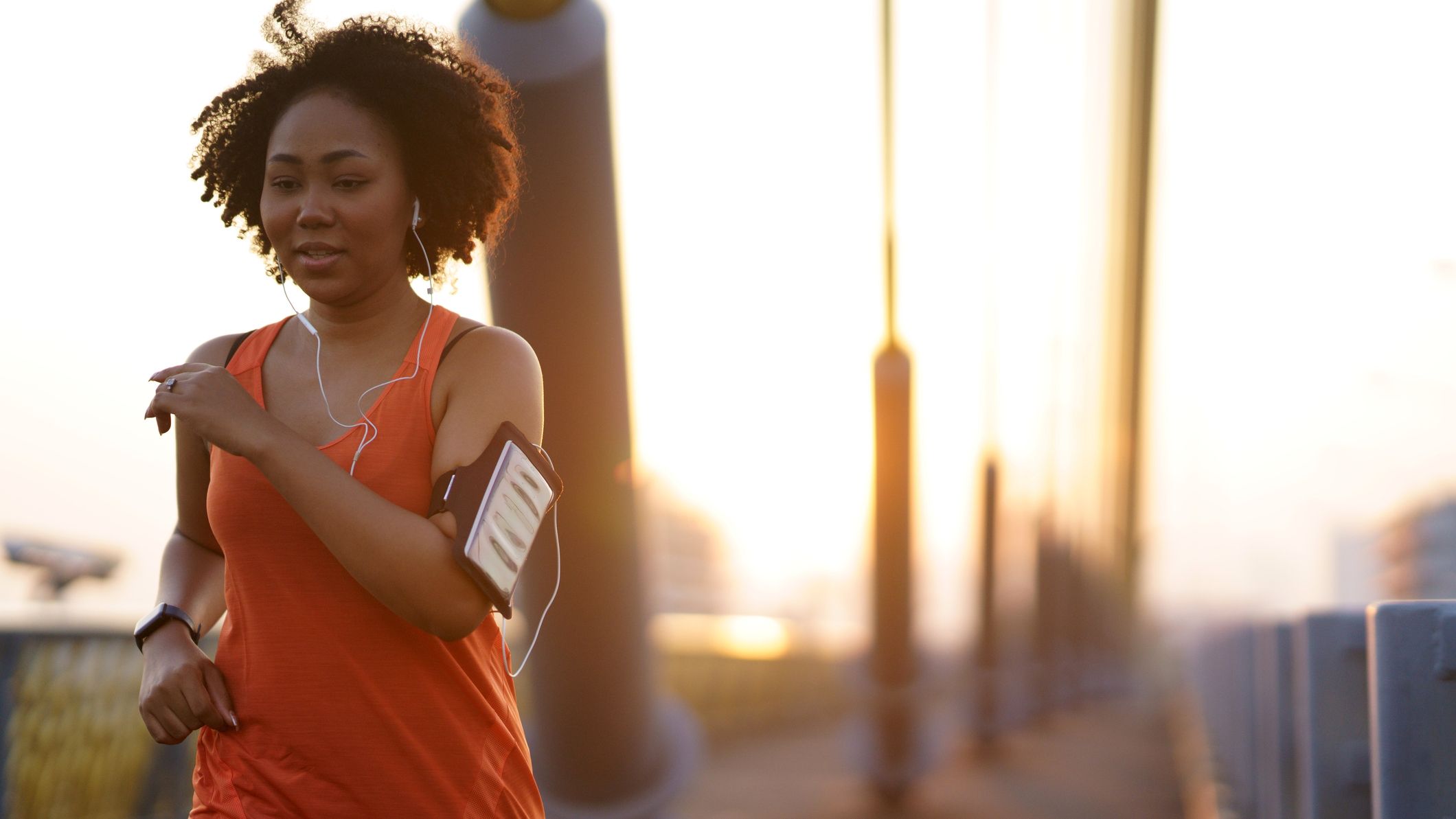 Woman power walks outdoors, wearing headphones and a phone in a phone holder on her arm