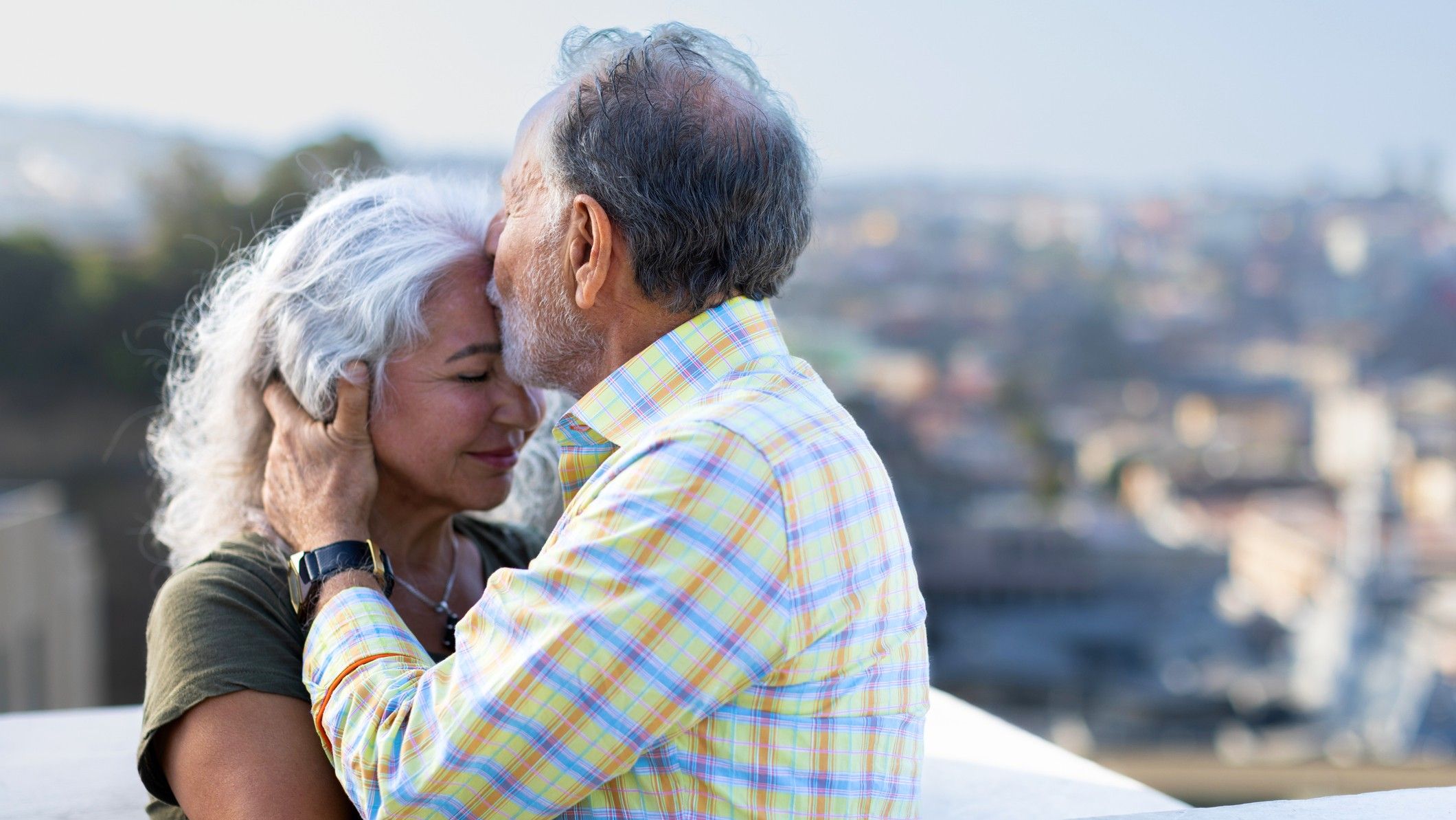An older couple is embracing, and he is kissing the woman's forehead tenderly.