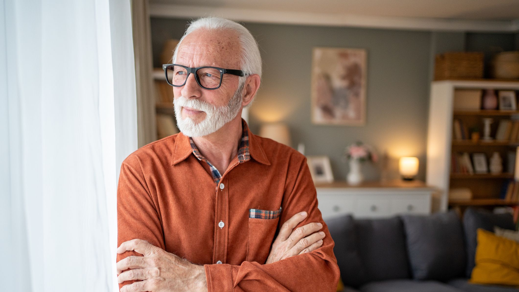Thoughtful senior man standing by the window in a cozy living room, arms crossed and gazing pensively at the view, reflecting on life&amp;amp;apos;s journey and cherished memories