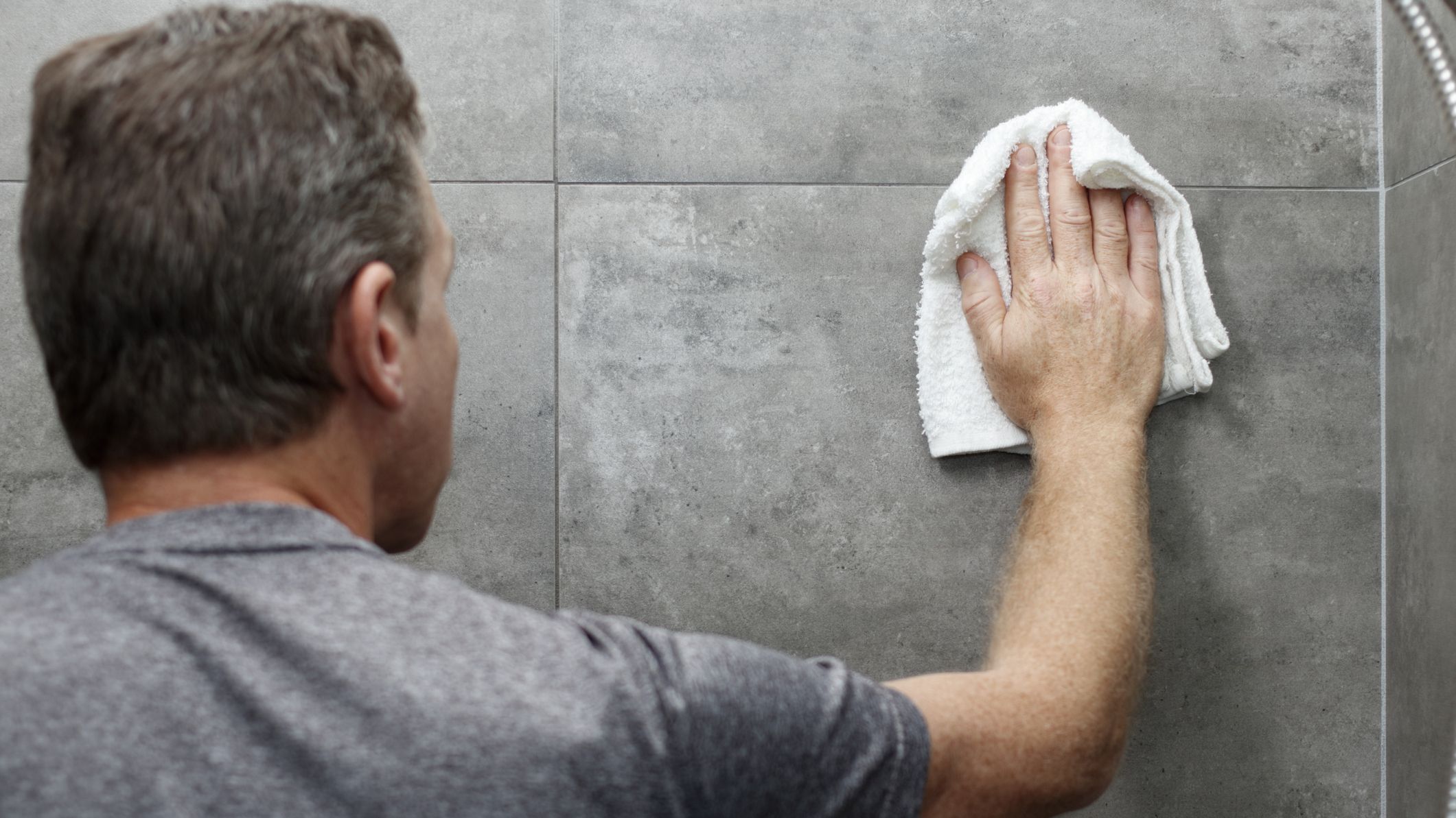 A man wiping down tile walls in bathroom with a dry rag.