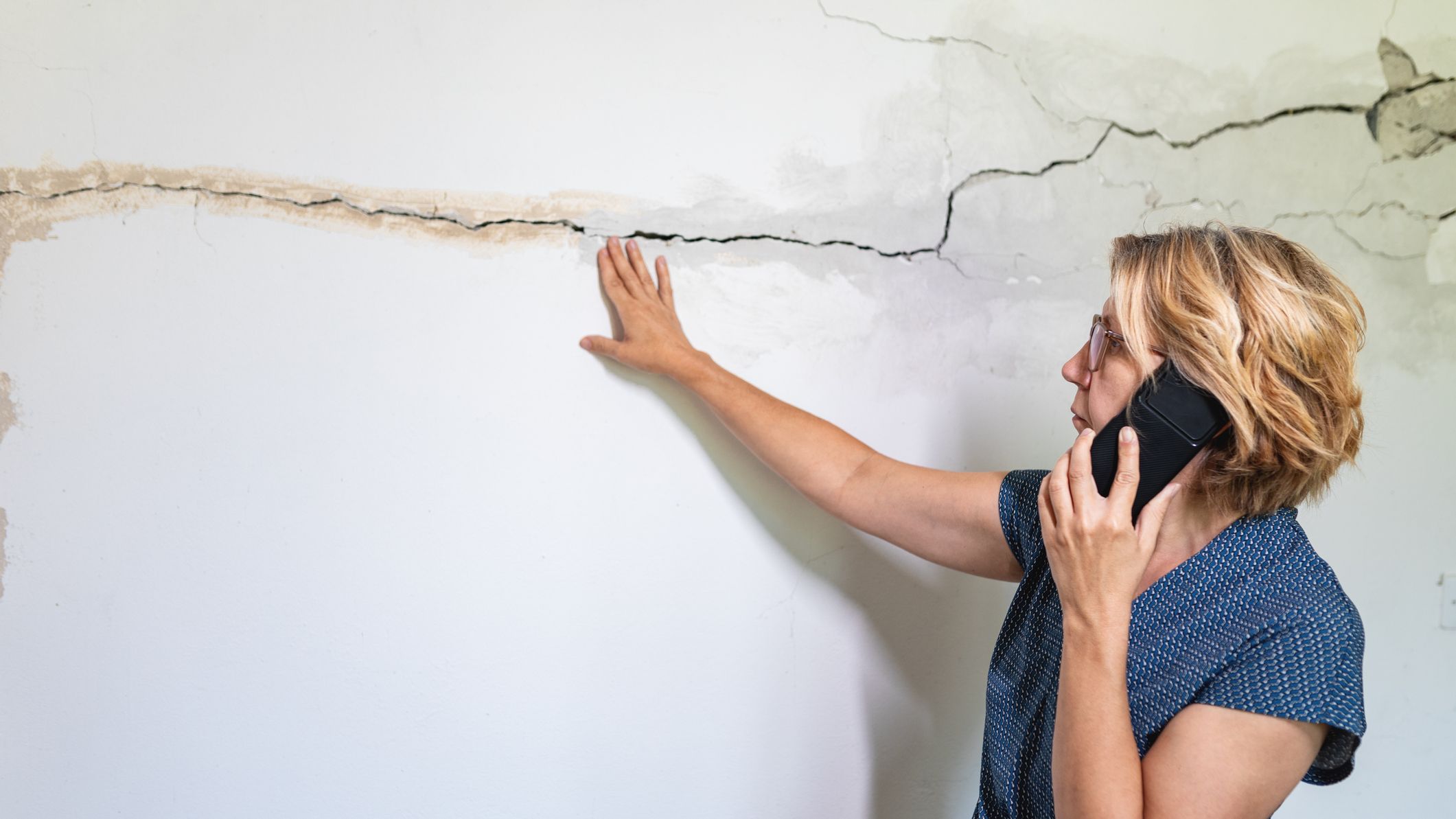 A woman on the phone while inspecting cracked wall.