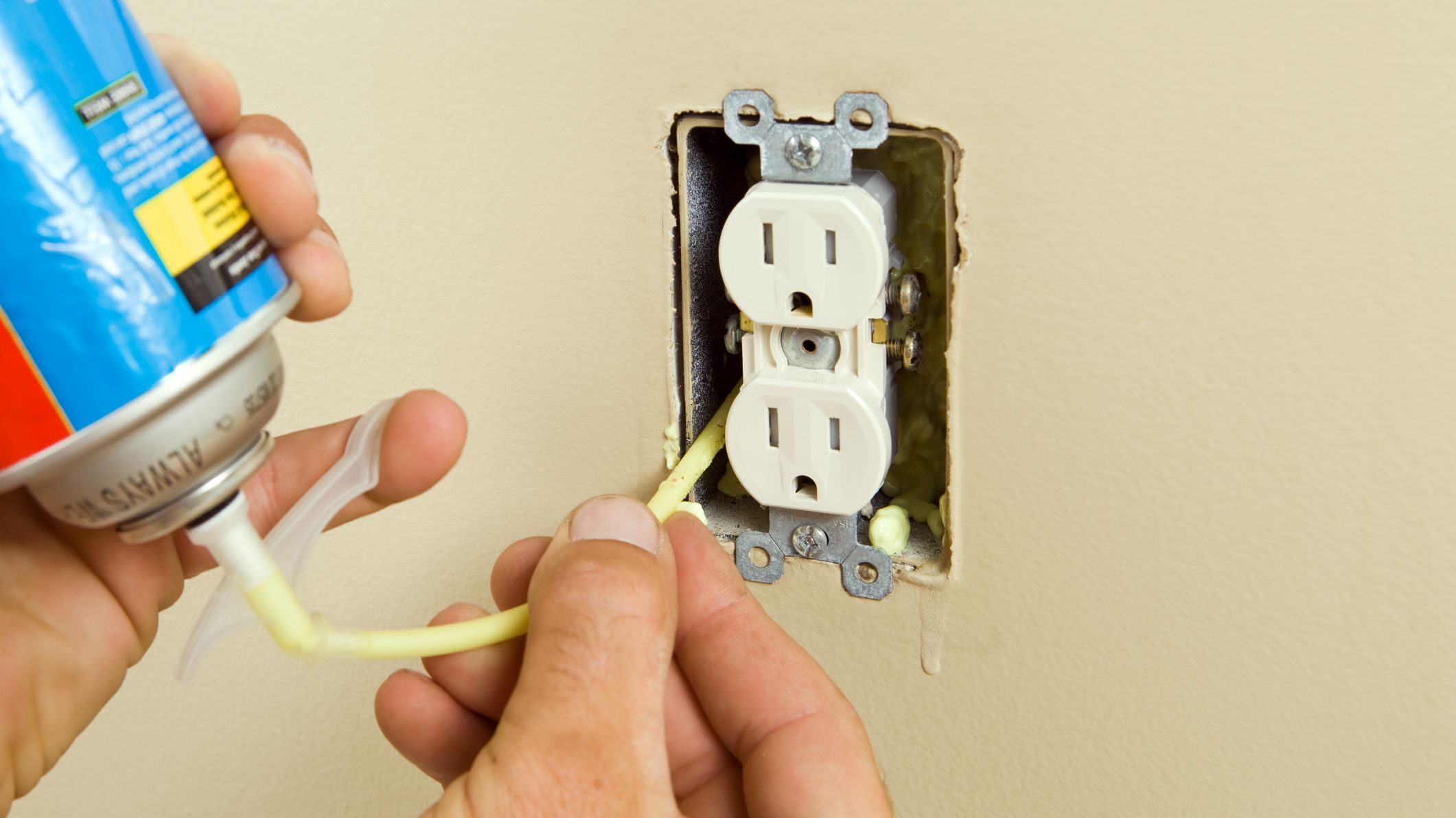 A hands holds a can of expandable foam insulation to insert it into the back of a wall outlet.