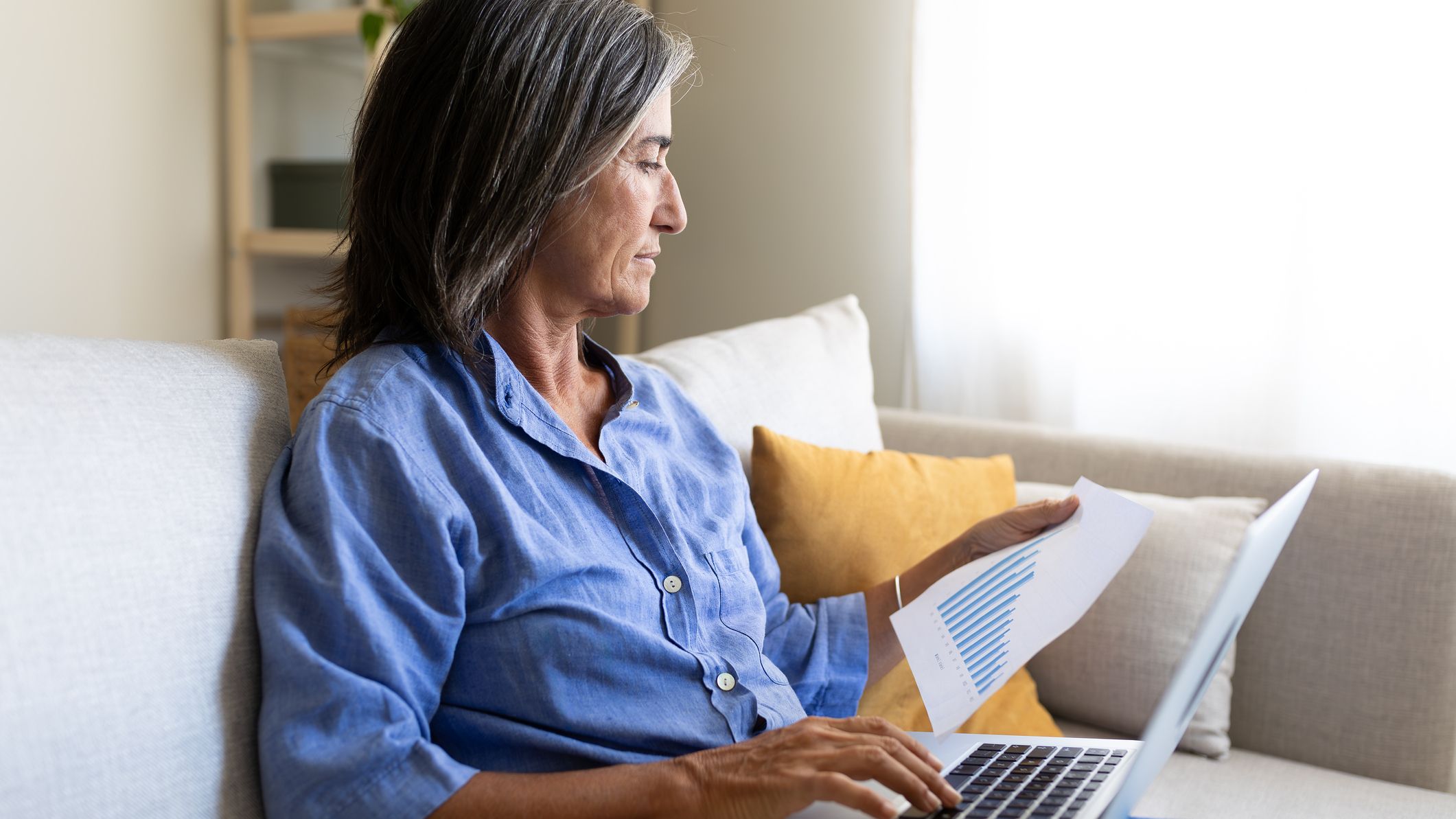 Mature businesswoman sitting on sofa using laptop and analyzing printed charts, working remotely from home