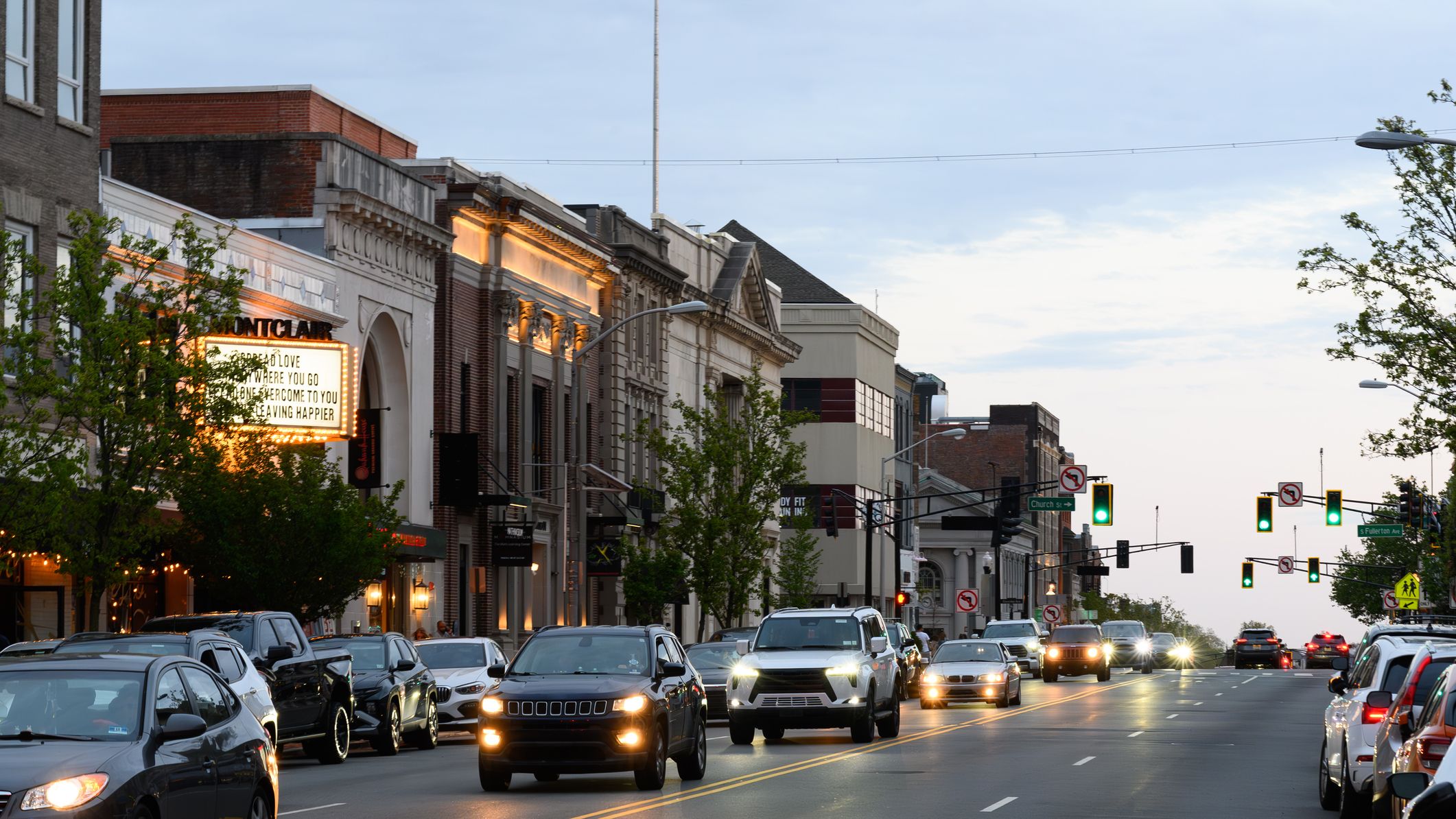 Cars fill the road in Montclair, New Jersey, moving through a downtown area lined with historic brick buildings and local storefronts, including a theater with an illuminated marquee.