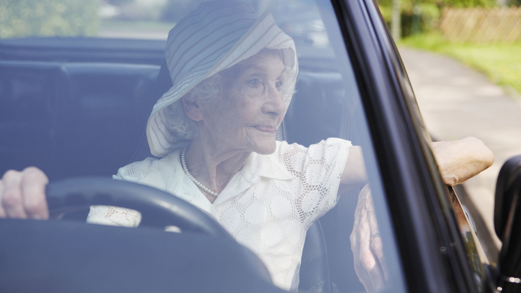 A woman in her eighties or nineties looks out the window from the driver's side of a car. She is wearing a hat and pearls.