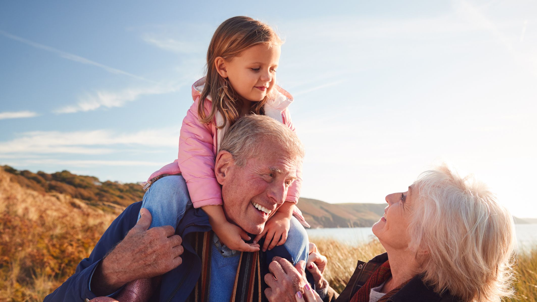 Grandfather Giving Granddaughter Ride On Shoulders As They Walk Through Sand Dunes With Grandmother.