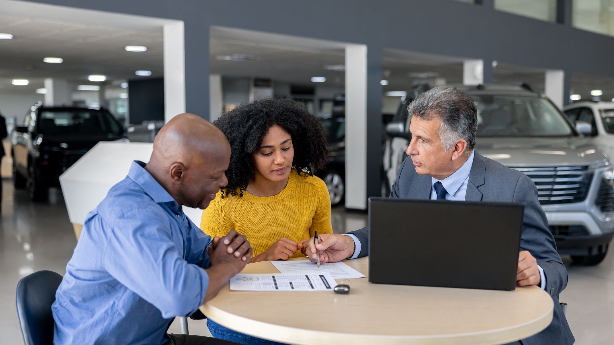 A car salesman discusses a contract with potential buyers while seated at a table inside a dealership.