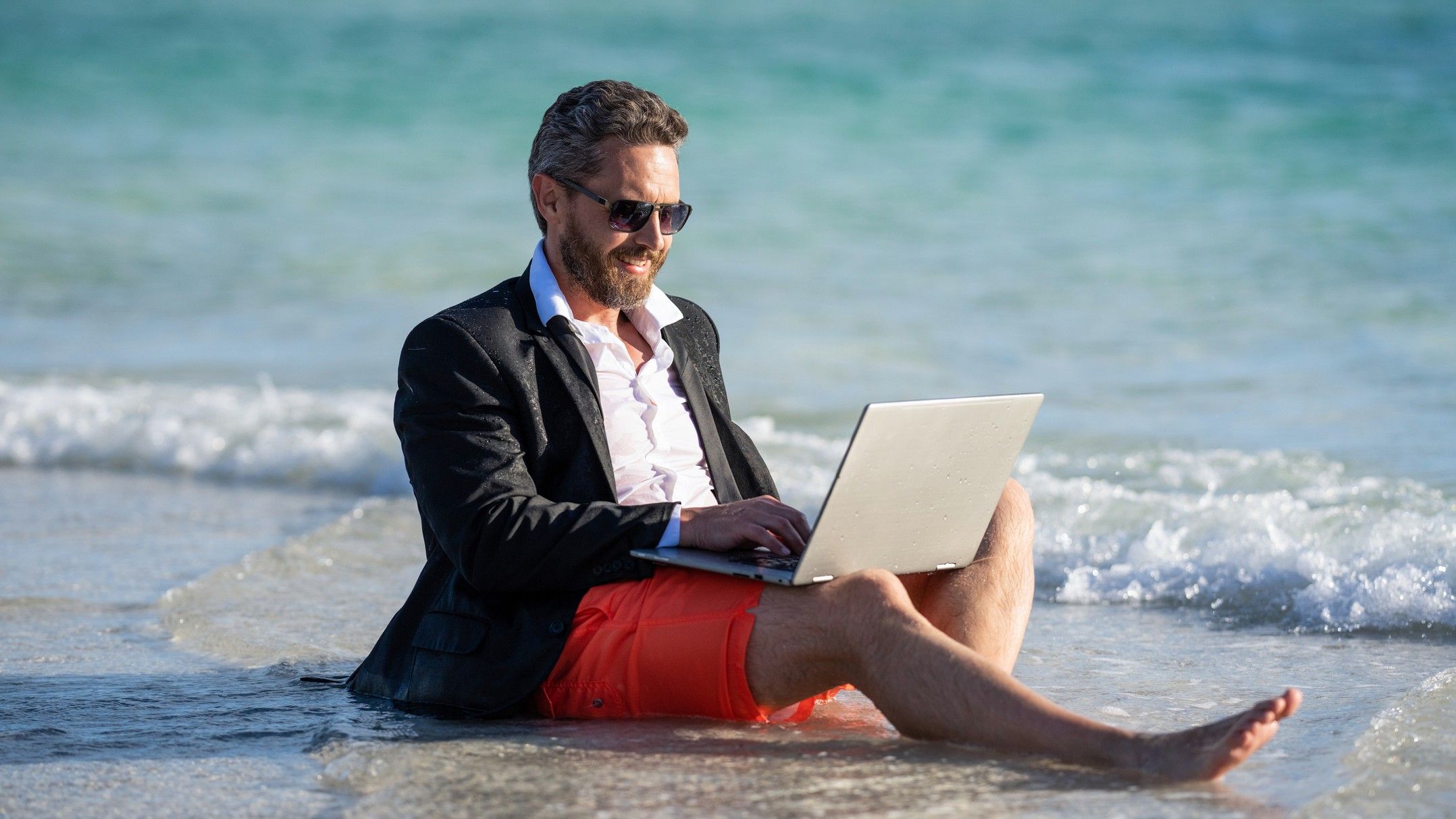 An attractive business man wearing a suit on his top half and a bathing suit on his lower half, lounges at the water line at the beach while on his laptop.