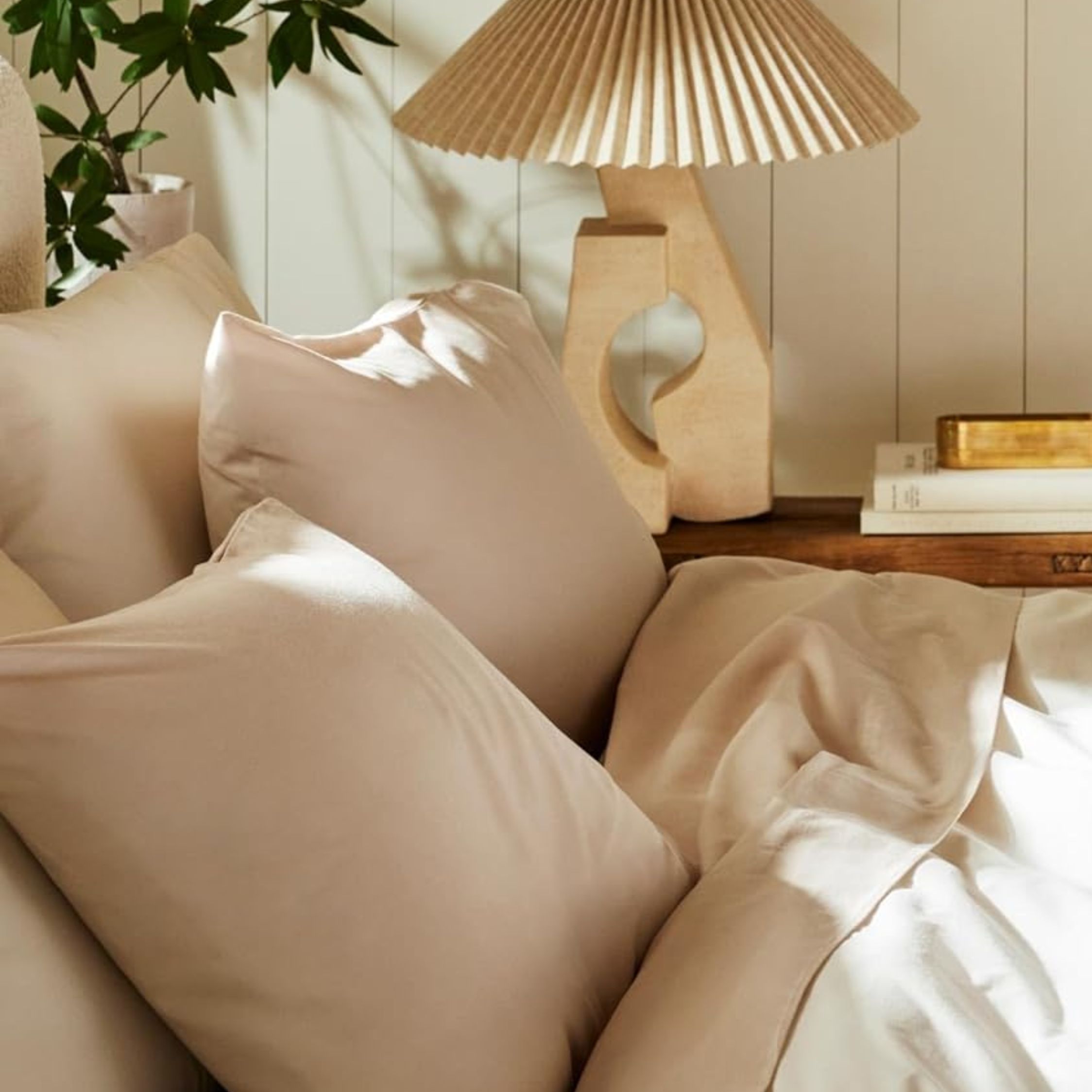 Beige Brooklinen bedding styled in a sunlit neutral bedroom that features a pleated table lamp and stack of books