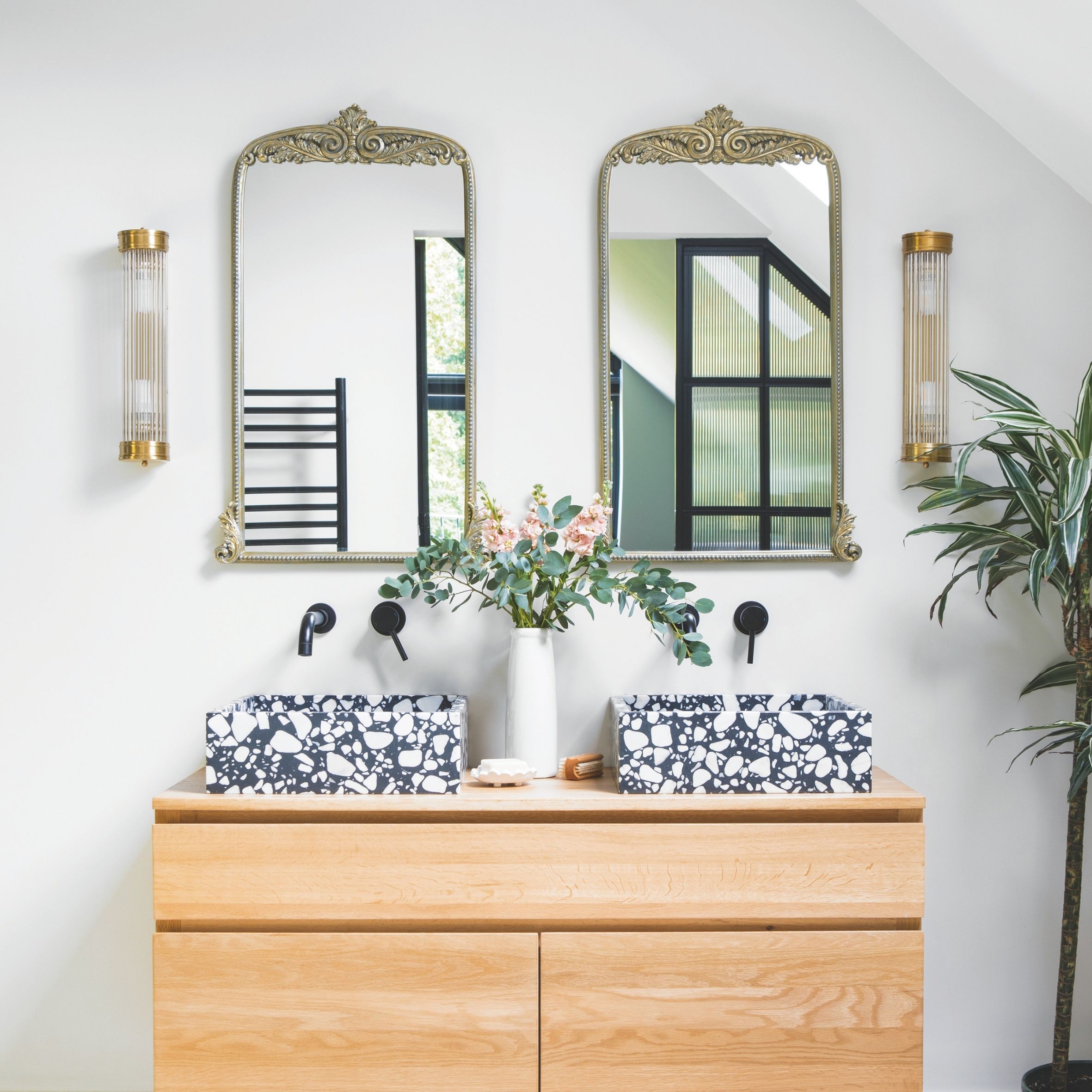 Double bathroom with wooden unit, 2 marble sinks and 4 black taps, 2 silver ornate mirrors and gold lighting either side