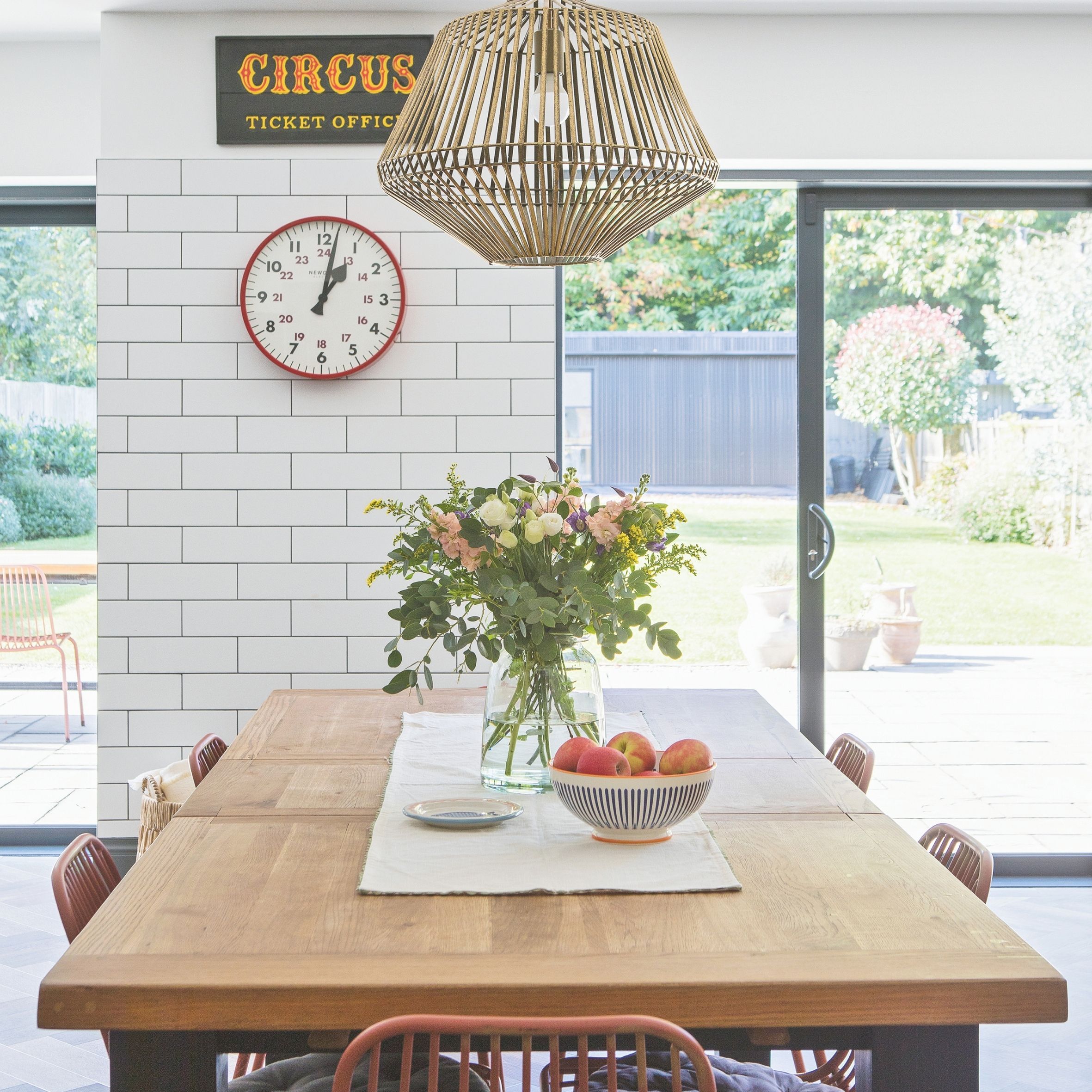 Dining room with white metro tiles on wall, red clock and wooden table. Red chairs and large bouquet in the middle of the table with fruit bowl