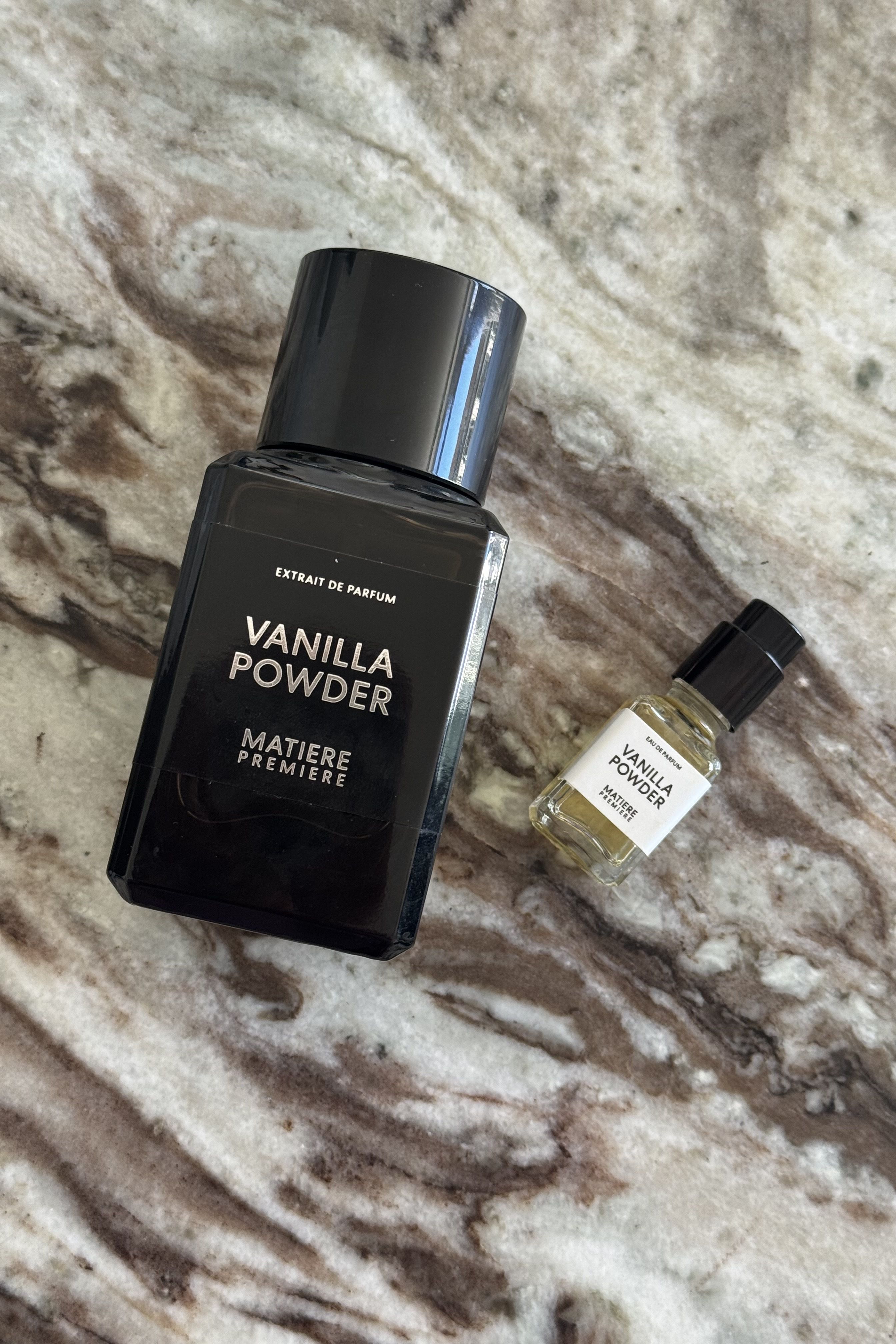 A bottle of Vanilla Powder Extrait and a travel-sized bottle of the eau de parfum on a marble textured background